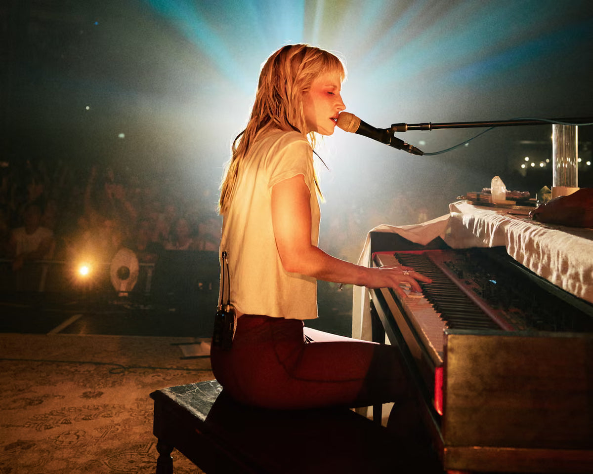 Hayley Williams sits at a piano on a stage, singing into a microphone. She is wearing a light beige short-sleeve top and dark red pants.